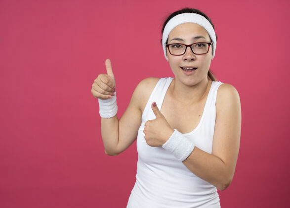 光学这位戴着眼镜 戴着头带和腕带的年轻运动女性给人留下了深刻的印象：两只手竖起大拇指 孤立在粉红色的墙上女孩腕带人