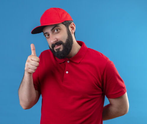 请年轻帅气的送货员穿着红色制服 戴着帽子 竖起大拇指 手放在蓝色的墙上人保持穿