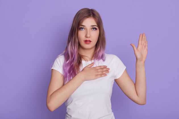 胸脯年轻专注的女性直视镜头 一只手放在胸前 另一只手举在胸前 展示手掌 咒骂 孤立在紫色的墙壁上女人心女士