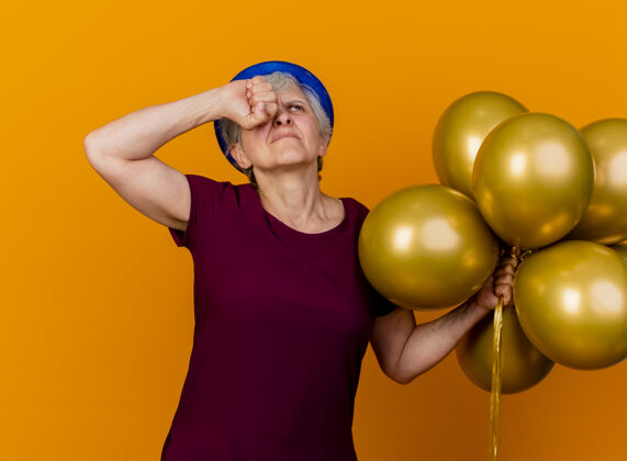 不愉快戴着派对帽的老妇人拿着氦气球 把拳头放在眼睛上 孤立地看着橘黄色的墙上 还有复制空间帽子拳头女人