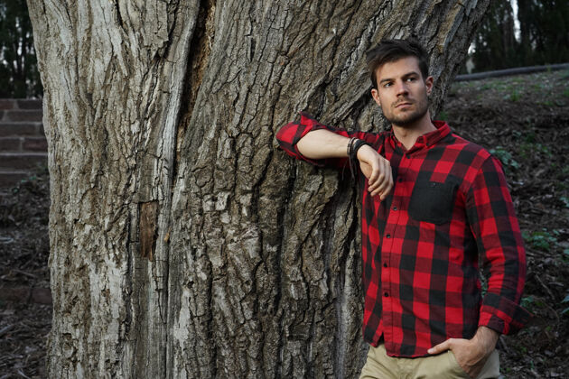 肖像一个迷人的年轻人靠在一棵老树上用手横拍瘦格子衬衫