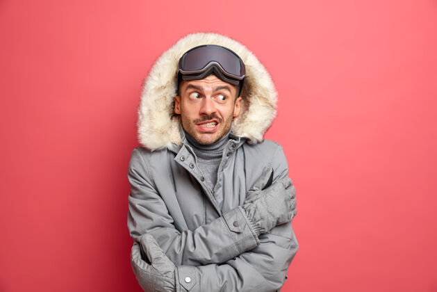 白种人英俊 困惑 没刮胡子的男子滑雪者在寒冷的霜天畏缩发抖 讨厌冬天寒冷 戴着滑雪板护目镜和灰色夹克寒冷冬季年轻