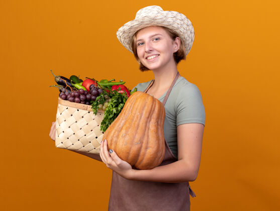 雌微笑着的年轻的斯拉夫女园丁戴着园艺帽 手里拿着蔬菜篮子和橘子上的南瓜南瓜篮子帽子