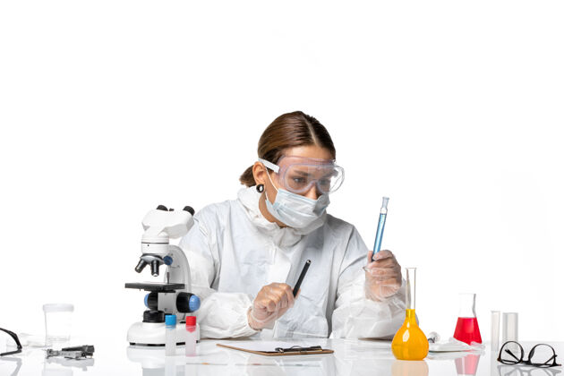 药品正面图：女医生穿着防护服 戴着口罩 拿着装有蓝色溶液的烧瓶 白色办公桌上有冠状病毒大流行医疗办公桌烧瓶