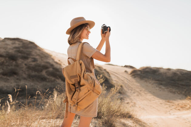 沙丘沙漠中穿着卡其色长裙的迷人时尚年轻女子 在非洲旅行 戴着帽子和背包 用老式相机拍照旅游美丽旅行者