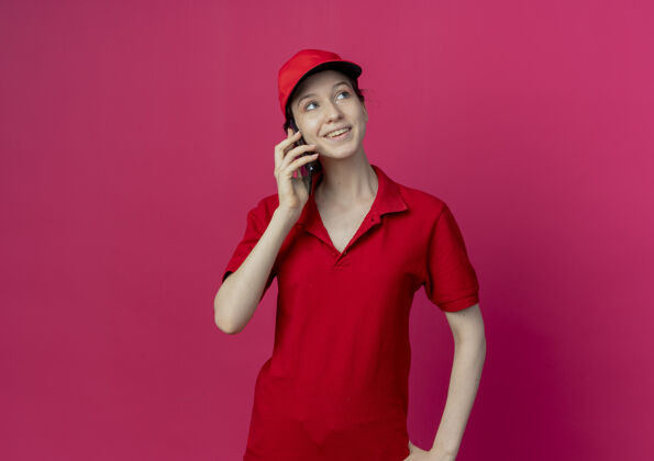 红色微笑着的年轻漂亮的送货女孩 穿着红色制服 戴着帽子 一边打电话 一边看着被隔离在深红色背景下的复制空间女孩制服帽子