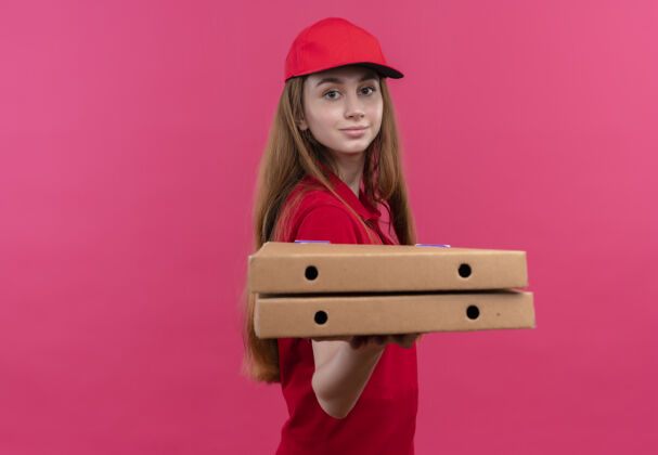 红色自信的年轻送货女孩穿着红色制服 伸出包裹 站在隔离的粉色空间和复印空间的侧视图中年轻自信粉色