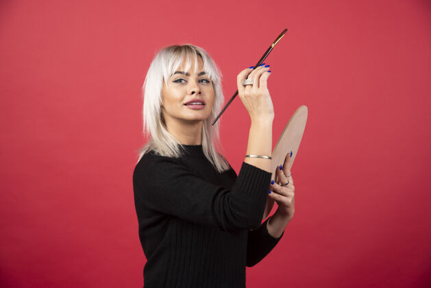 女人年轻的女艺术家拿着艺术用品站在红墙上调色板画笔画笔