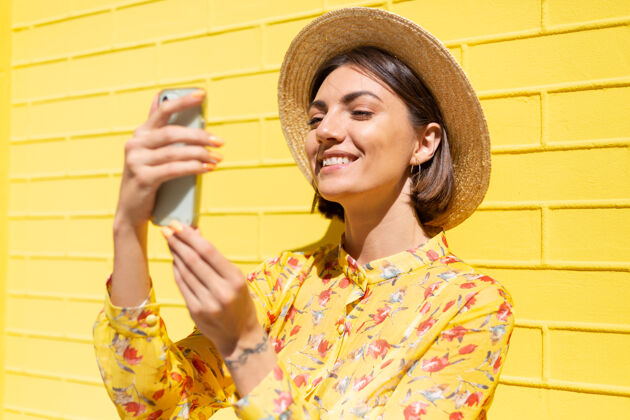 欢呼穿着黄色夏装戴着黄色帽子的女人在黄色的砖墙上冷静而积极地拿着手机相机成人墙