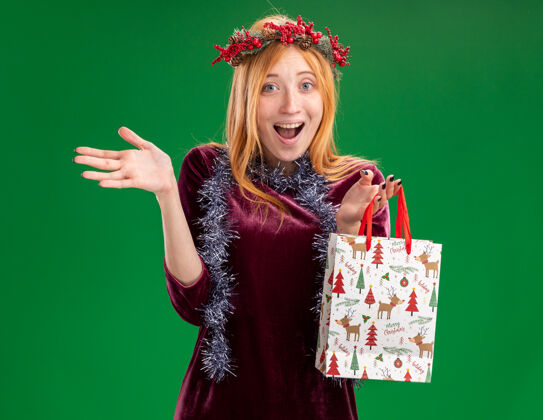 手兴奋的年轻漂亮的女孩穿着红色的衣服 脖子上戴着花环和花环 手里拿着礼品袋 手被隔离在绿色的背景上脖子红色圣诞快乐