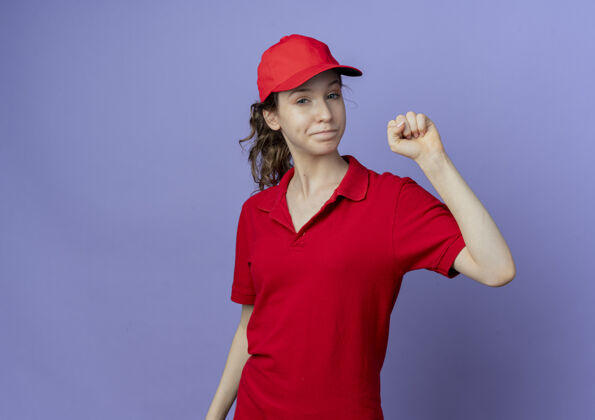 手势自信的年轻漂亮的送货女孩穿着红色制服 戴着帽子 在紫色背景上显示出举起的拳头姿势 并留有复制空间红色背景提高