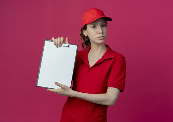 女孩未出租的年轻漂亮的送货女孩在红色制服和帽子显示剪贴板在深红色背景与复制空间隔离显示不愉快制服