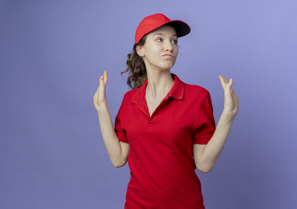 手自信的年轻漂亮的送货女孩穿着红色制服 戴着帽子 一边看一边显示空的手 紫色背景上与复制空间隔离信心空背景