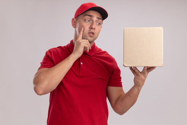 递送年轻的送货员穿着制服 戴着帽子 看着隔离在白墙上的箱子人盒子人