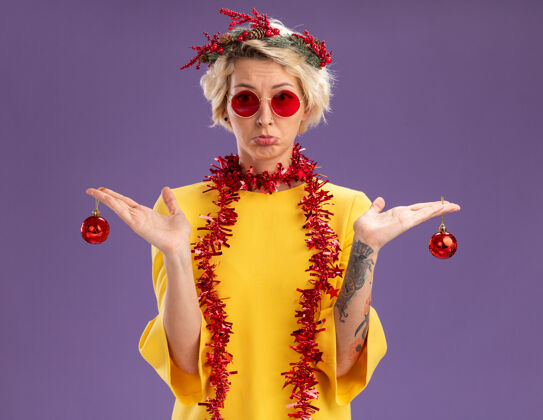 年轻笨手笨脚的金发女郎戴着圣诞花环 脖子上戴着金属丝花环 戴着眼镜 手里拿着圣诞饰品 看着紫色背景上孤立的摄像机女人花环紫色