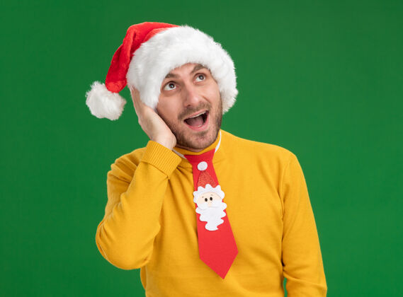 领带印象深刻的年轻白种人戴着圣诞帽 打着领带 手放在头上看着绿色背景上孤立的一面帽子头男人
