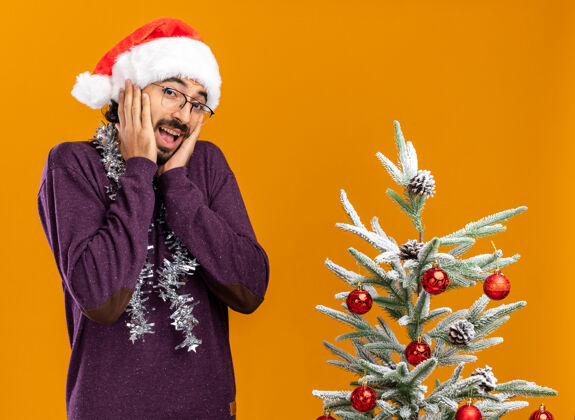 圣诞快乐兴奋的年轻帅哥站在圣诞树旁 戴着圣诞帽 脖子上戴着花环 手放在脸颊上 橘黄色的背景隔离开来手脸颊兴奋