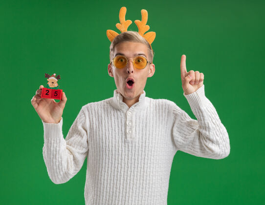 年轻令人印象深刻的年轻帅哥戴着驯鹿鹿角头带戴着眼镜拿着圣诞树玩具和日期看着相机指着绿色背景上孤立绿色指向玩具