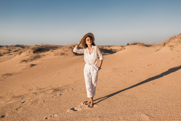 体验一个穿着白色衣服 戴着草帽 在沙漠沙滩上行走的时尚美女旅游女性模特