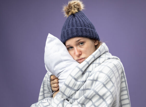 围巾悲伤的生病的年轻女孩穿着白色长袍 戴着冬天的帽子 围巾裹着格子呢 抱枕孤立地躺在紫色的床上悲伤枕头长袍