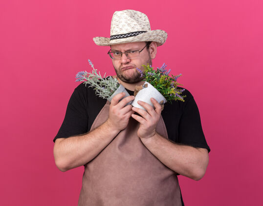 花园贪婪的年轻男园丁戴着园艺帽 手里拿着孤立在粉红色墙上的花盆里的花贪婪园艺帽子