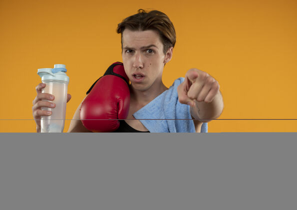 肩膀自信的年轻运动型男人 肩上戴着毛巾和拳击手套 拿着水瓶 在橙色的墙上向你们展示孤立的姿势年轻拳击手势