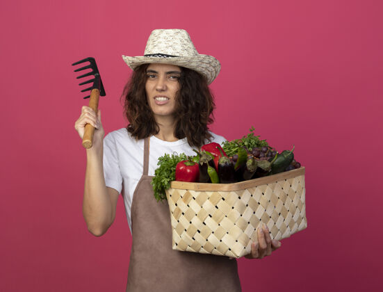 帽子自信的年轻女园丁穿着制服戴着园艺帽拿着菜篮用耙子篮子女人花园