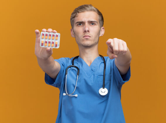 制服年轻严格的男医生 穿着医生制服 手持听诊器 在橙色的墙上显示你的手势听诊器握着药丸