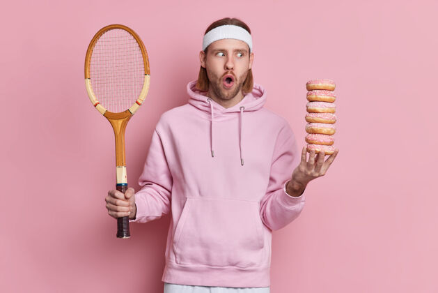 男士震惊的白种人脸上带着惊愕的表情 在网球比赛中摔了一跤 手里拿着球拍和一堆甜甜圈过着积极的生活技能男士印象