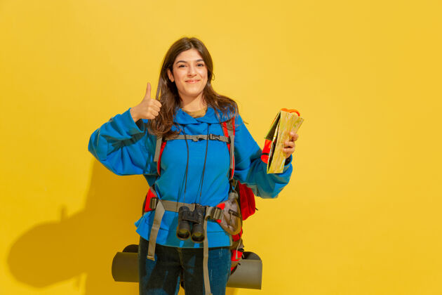 肖像微笑着 竖起大拇指一个快乐的年轻高加索旅游女孩的肖像 带着包和望远镜 隔离在黄色的工作室背景上准备旅游度假胜地 人类的情感 度假衣服年轻指向