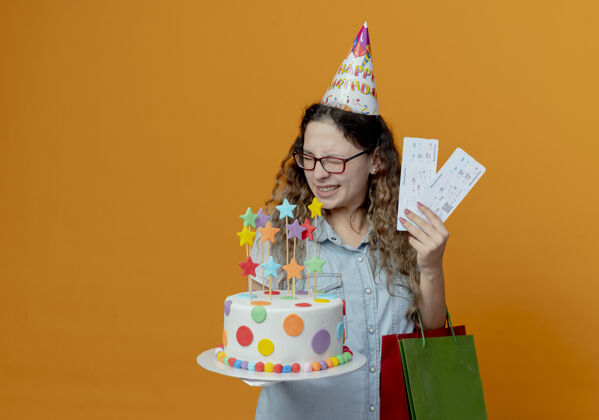 帽子闭上眼睛 戴着眼镜 戴着生日帽 拿着门票和生日蛋糕的快乐年轻女孩 礼物袋孤立在橙色背景上举行蛋糕年轻