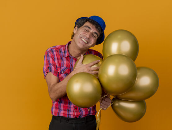 表情快乐的中年派对男戴着派对帽拿着气球闭着眼睛孤立在橘黄色的墙上有复制空间闭上人快乐