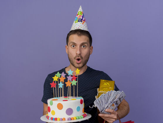 表情戴着生日帽的帅哥拿着生日蛋糕礼盒和钱 隔离在紫色的墙上 留着复制空间生日礼物钱