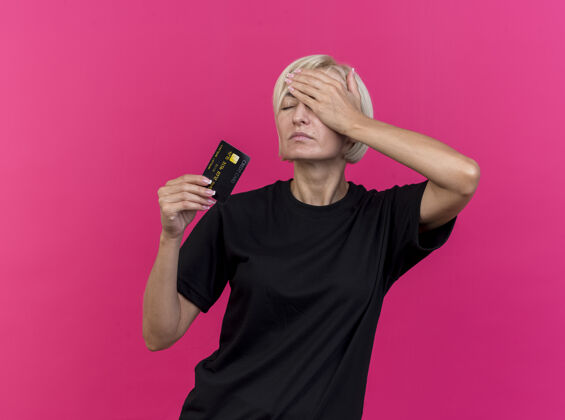 人疼痛的中年金发斯拉夫妇女拿着信用卡 手放在头上 闭着眼睛 隔离在粉红色的墙上 还有复印空间感情脸女人