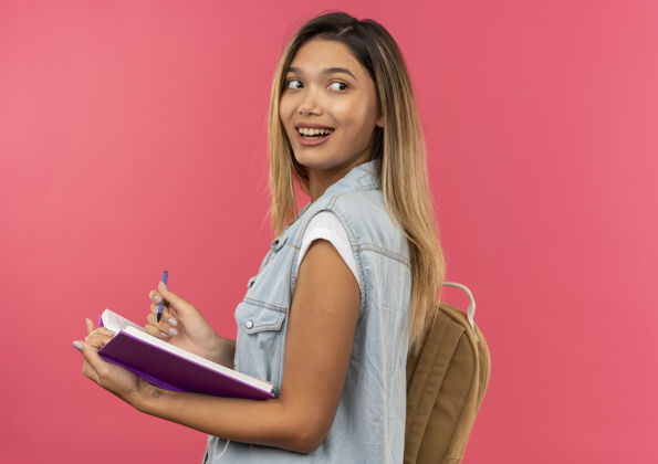 背部快乐的年轻漂亮的学生女孩戴着背袋站在侧视图中 拿着打开的书和笔 看着后面孤立的粉红色查看年轻立场