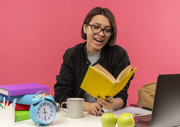 年轻戴着眼镜坐在书桌旁拿着书做作业的快乐的年轻女学生学生拿着阅读
