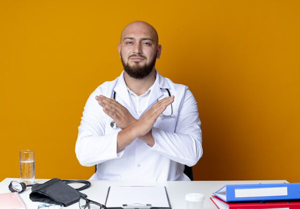 手势年轻的秃头男医生穿着医用长袍和听诊器坐在工作台上 手持医疗工具 在橙色背景上显示出不孤立的姿态医疗医生长袍