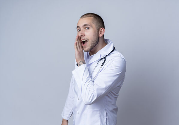 年轻令人印象深刻的年轻男医生穿着医用长袍 听诊器挂在脖子上 用手在嘴旁低语 隔离在白色皮肤上脖子靠近口腔