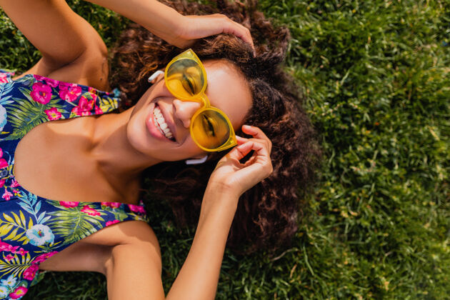 年轻年轻时尚的黑人女子 戴着无线耳机听音乐 躺在公园的草地上玩得很开心 夏日的时尚风格 五颜六色的时髦装扮 俯瞰风景酷黄色明亮