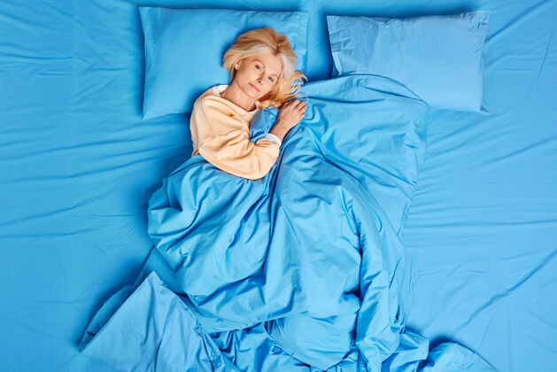 顶部冷静的欧洲中年妇女看到好梦后醒来心满意足摆出好姿势睡在蓝色毯子下穿着睡衣感觉舒服享受懒散的一天床上时间和舒适的早晨概念欧洲年轻休息
