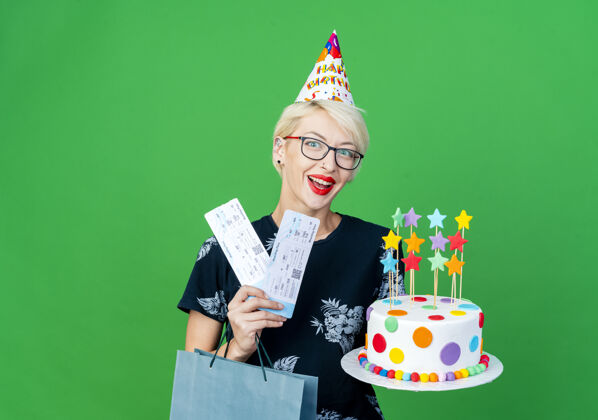 蛋糕令人印象深刻的年轻金发派对女孩戴着眼镜和生日帽拿着生日蛋糕明星机票和纸袋看着相机隔离在绿色背景与复制空间印象持有帽子