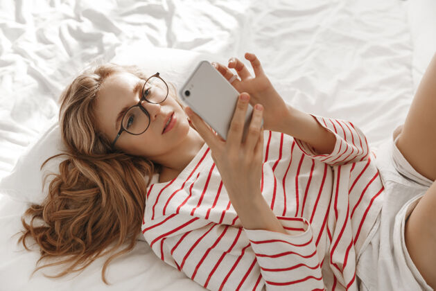 电话年轻的微笑的女人坐在家里的床上 用她的智能手机发短信年轻放松房子