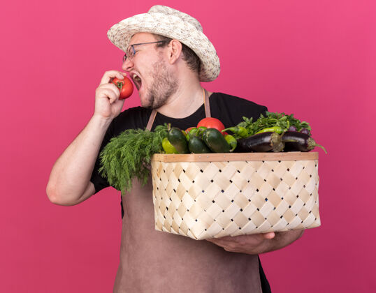 立场站在侧视图年轻的男园丁戴着园艺帽拿着蔬菜篮子在粉红色的墙上尝试西红柿隔离篮子帽子蔬菜