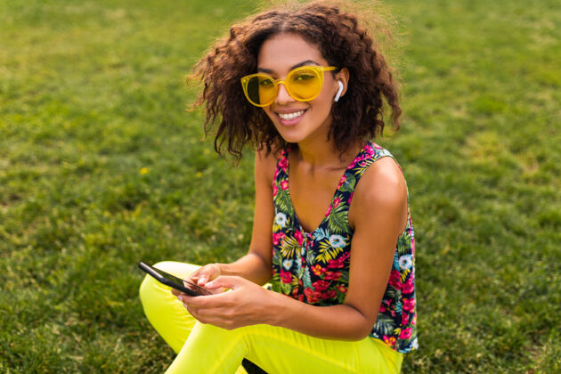 太阳镜年轻时尚微笑的黑人女子使用智能手机 通过无线耳机听音乐 在公园里玩得很开心享受多彩酷
