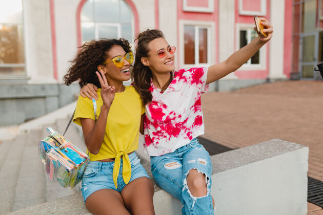 年轻快乐的年轻女孩朋友们微笑着坐在街上用手机自拍 女人们一起开心兴奋微笑时尚