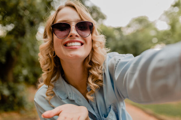 微笑迷人的金发微笑的女人穿着夏装在公园里散步 在电话里自拍有趣春天夏天