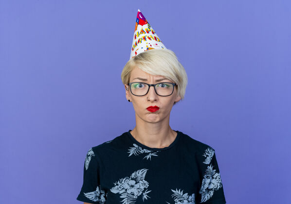 眼镜戴着眼镜 戴着生日帽 在紫色背景下看着孤立的相机的金发派对女孩年轻金发生日