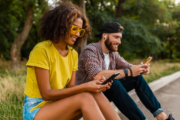 酷快乐的年轻微笑的朋友坐在公园里用智能手机 男人和女人一起玩电话女人时尚