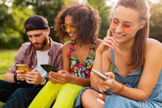 积极快乐的年轻朋友们坐在公园里用智能手机微笑 男人和女人一起玩时尚太阳镜兴奋