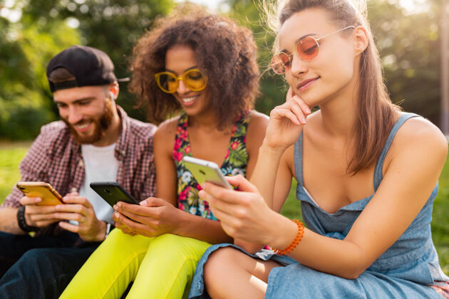 手机快乐的年轻朋友们坐在公园里用智能手机微笑 男人和女人一起玩年轻酷积极